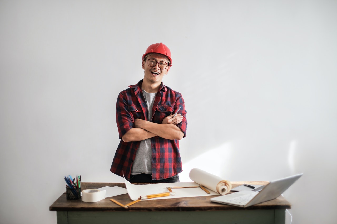 A man wearing a hard helmet and smiling beside a table with chart paper, plans, and a scale